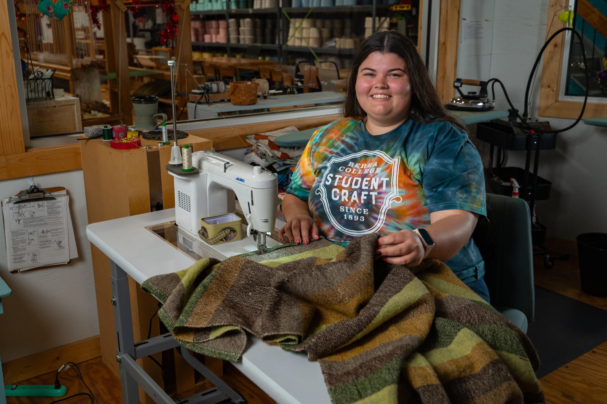 Berea student, Dani '25, smiling at the camera while holding a blanket that she's working on.